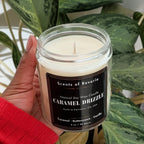 Hand holding a candle labeled 'Caramel Drizzle' in front of a potted plant