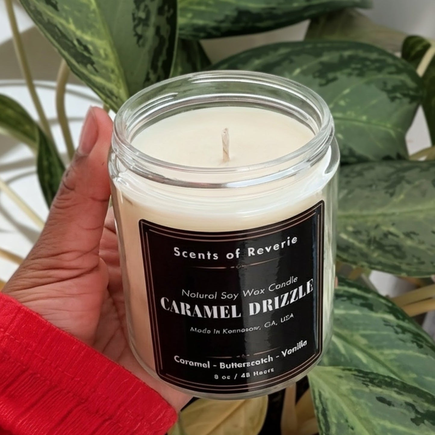 Hand holding a candle labeled 'Caramel Drizzle' in front of a potted plant