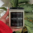 Hand holding a candle labeled 'Caramel Drizzle' in front of a plant