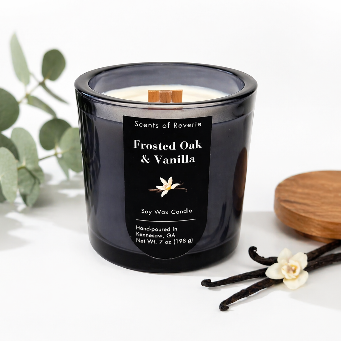 Black candle labeled 'Frosted Oak & Vanilla' with vanilla beans and a leaf on a white background