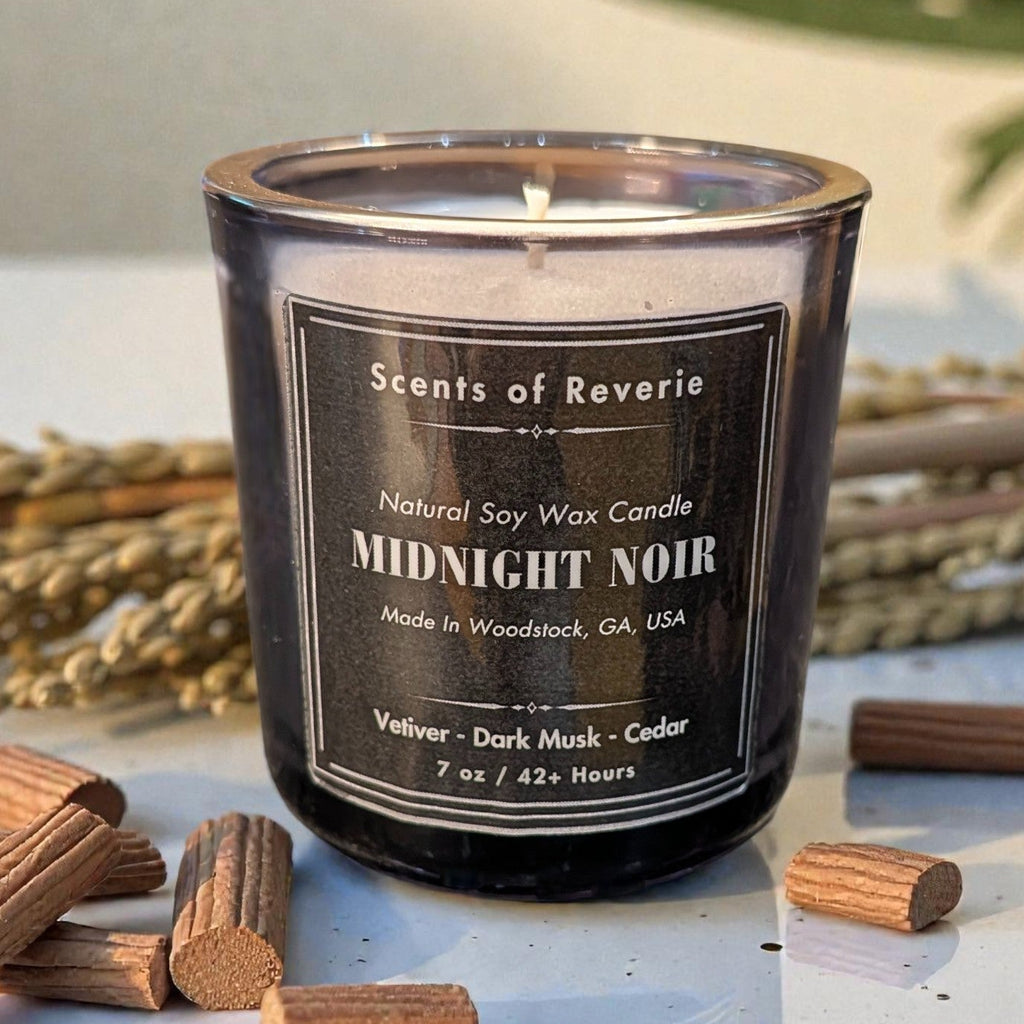 Candle labeled 'Scents of Reverie Midnight Noir' with wooden sticks and lavender on a light surface.