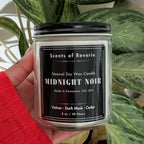 Candle labeled 'Midnight Noir' held in front of a green plant