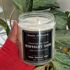 Person holding a 'Midnight Noir' candle in front of green plants