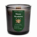 A soy wax candle in a smoky translucent dark glass jar with a dark green label reading 'Scents of Reverie Mossy Meadows' and a small wood wick visible at the top, photographed on a white background.