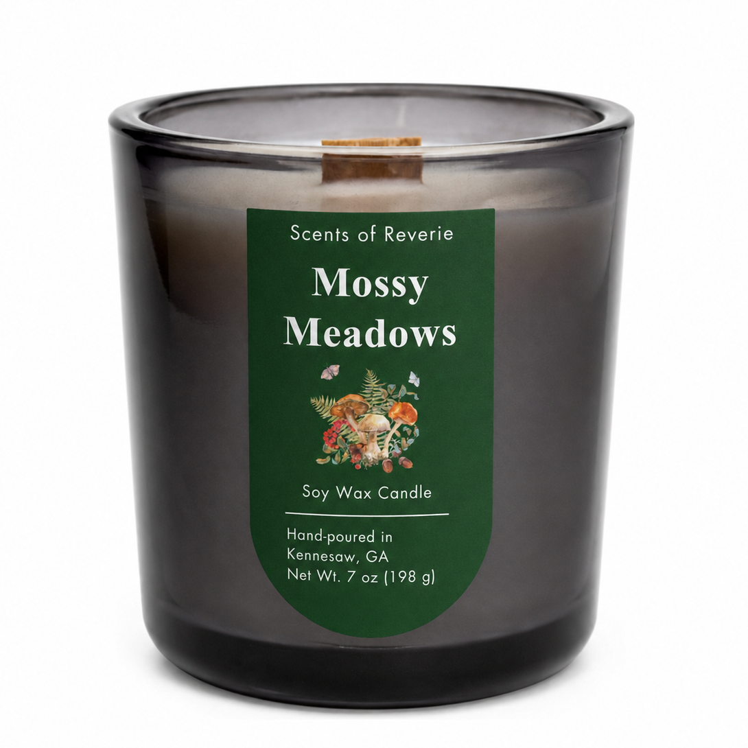A soy wax candle in a smoky translucent dark glass jar with a dark green label reading 'Scents of Reverie Mossy Meadows' and a small wood wick visible at the top, photographed on a white background.