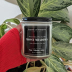 Candle labeled 'Mossy Meadows' held by a person in front of a plant