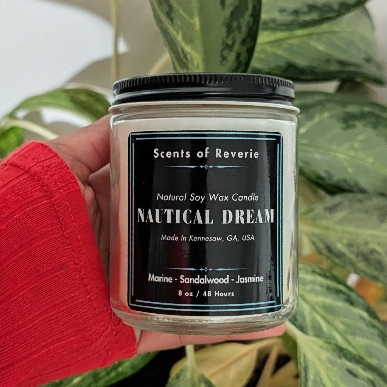 Candle labeled 'Nautical Dream' held in front of a potted plant