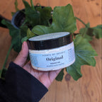 Hand holding a jar labeled 'Original' in front of a plant on a wooden floor.