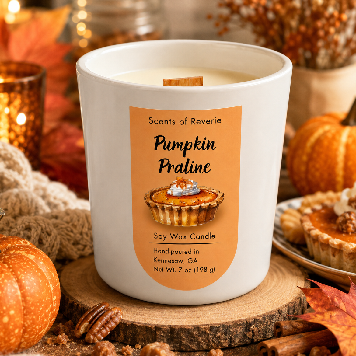 A white wax candle in a white glass jar with a warm orange label reading 'Pumpkin Praline' and 'Scents of Reverie,' styled on a wood slice with pumpkins, fall leaves, nuts, and a cozy autumn background.