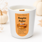 A white ceramic soy wax candle in a glass jar with a warm orange label reading 'Pumpkin Praline' and 'Scents of Reverie.' A small pumpkin, leaf, and dried floral decor are softly blurred in the background.