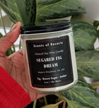 Hand holding a candle labeled 'Sugared Fig Dream' in front of a plant