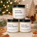 Three scented candles in glass jars are arranged in a holiday-themed setting. The top jar is labeled 'Smells Like.