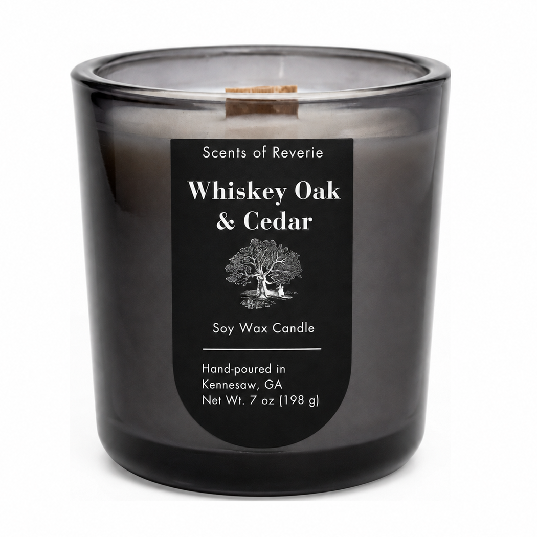 A 7 oz soy wax candle in a dark, translucent smoky gray glass jar with a black label reading 'Scents of Reverie Whiskey Oak & Cedar'.