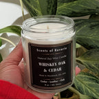 Hand holding a candle labeled 'Whiskey Oak & Cedar' with green plants in the background