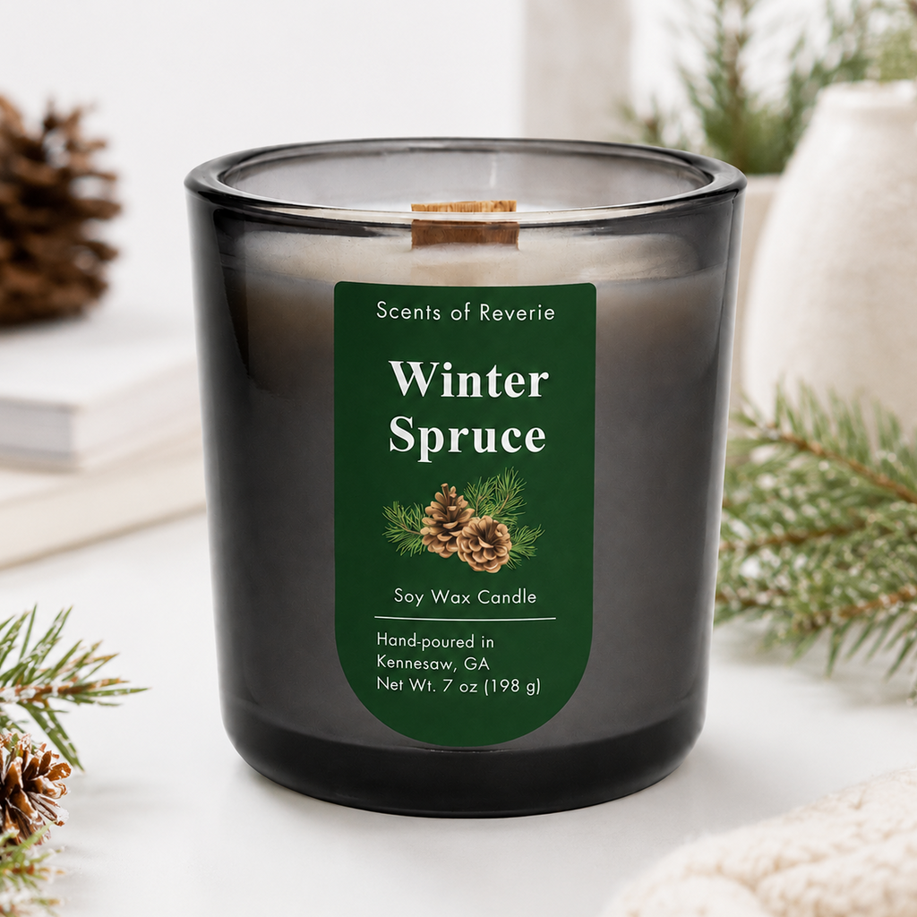 A dark gray glass soy wax candle with a green label reading 'Winter Spruce' by Scents of Reverie, styled on a white surface with pine branches, a pinecone, books, and a cream vase in the background.