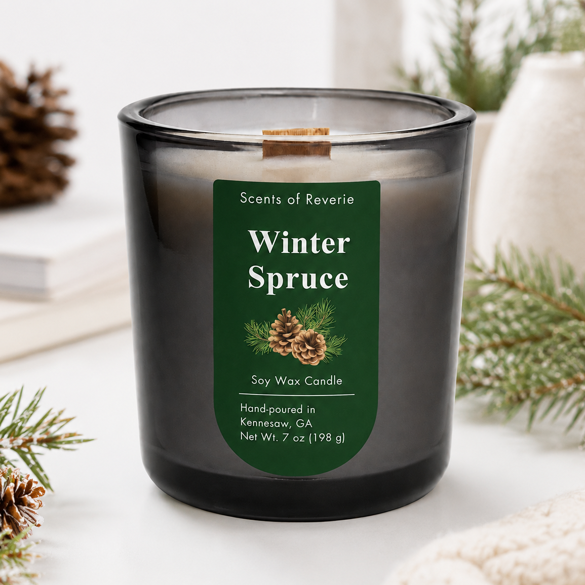 A dark gray glass soy wax candle with a green label reading 'Winter Spruce' by Scents of Reverie, styled on a white surface with pine branches, a pinecone, books, and a cream vase in the background.