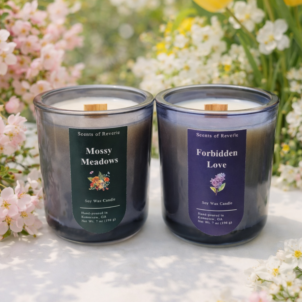 Two candles labeled 'Mosy Meadows' and 'Forbidden Love' on a surface with flowers in the background.
