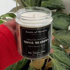 Candle labeled 'Scents of Reverie Mossy Meadows' held in front of a green plant.
